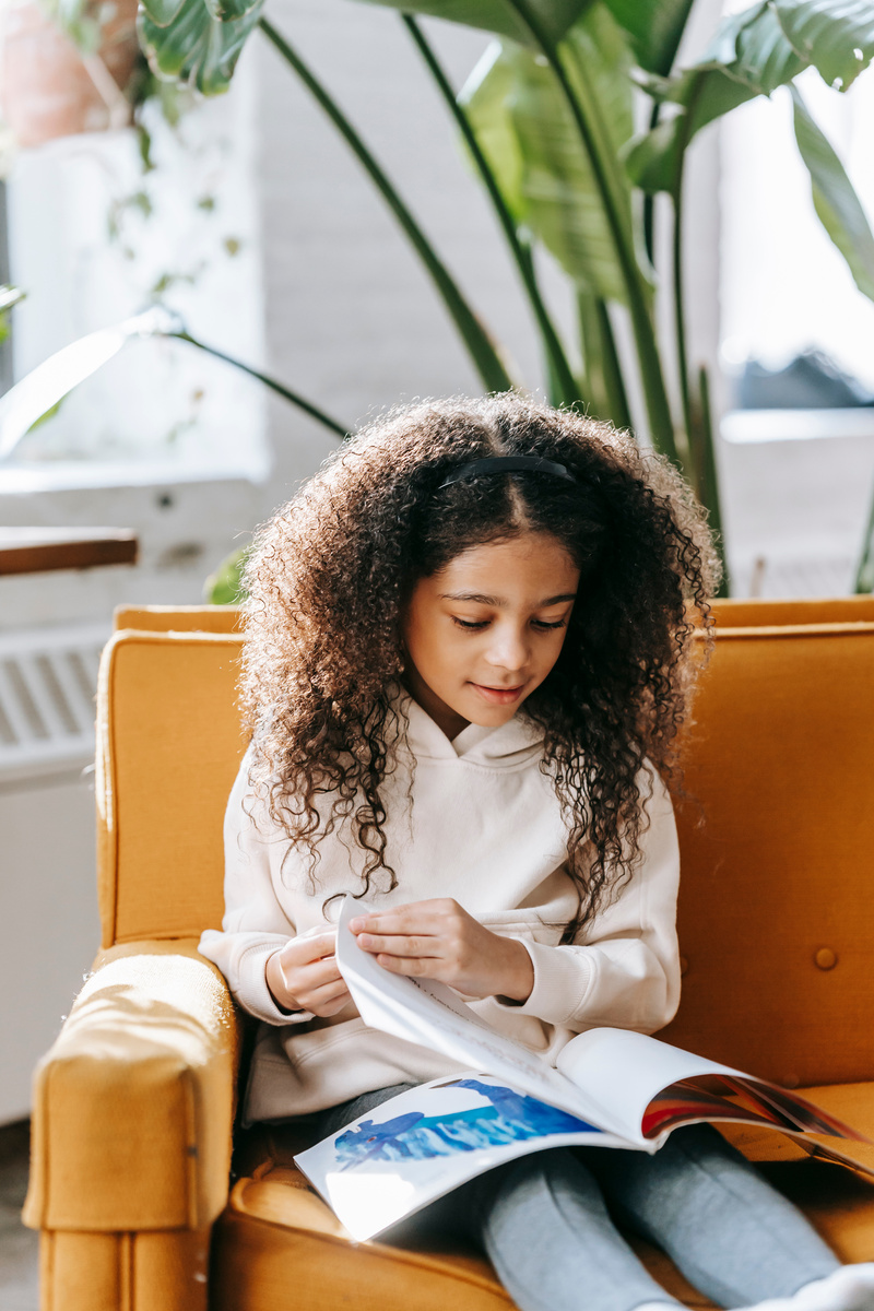 Pensive black girl reading magazine in living room in sunny day